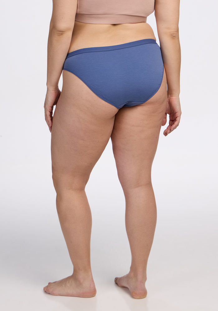 A woman stands barefoot, facing away from the camera, wearing a beige crop top and Woolx Roxie Bikini Underwear in Skyline blue against a plain light background.