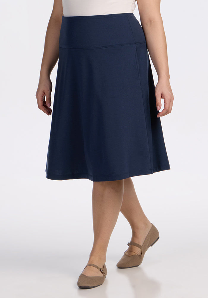 A person wearing the Woolx Marilyn Flowy Midi Skirt in Deep Navy, paired with a white top and tan flats, stands against a plain white background. 
