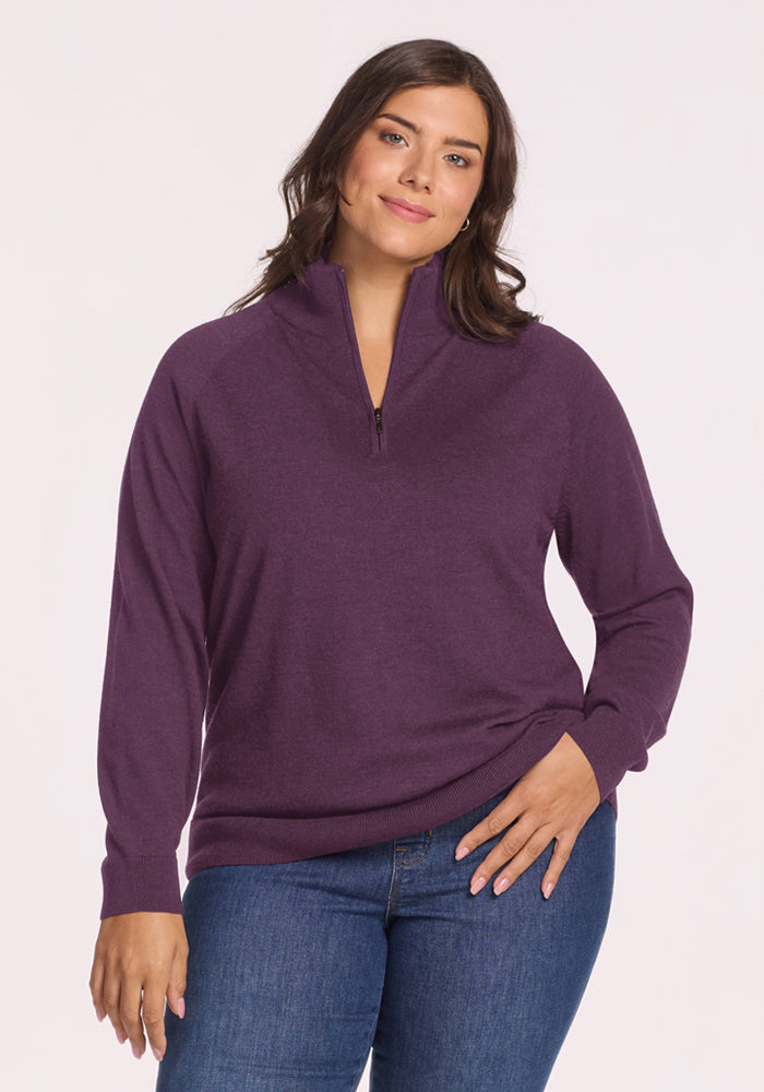 Smiling, a woman wears the Woolx Collins Half Zip Sweater in Purple Iris and blue jeans against a white background. 