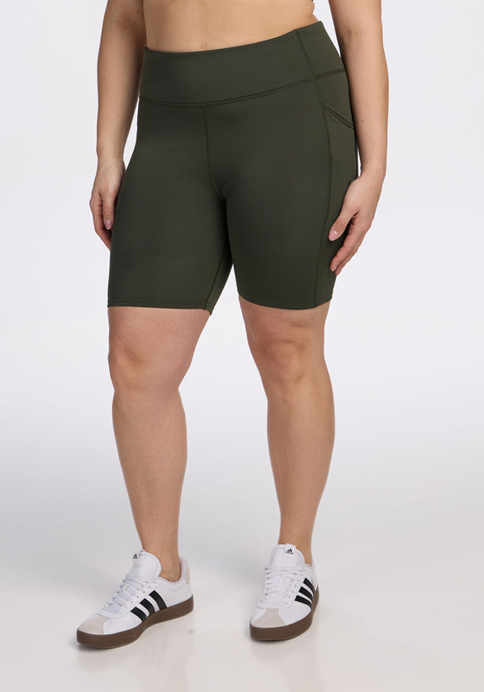 A person wearing Woolx Merino Flex® Dani Bike Shorts in Everleaf and white Adidas sneakers with black stripes stands against a plain white background, with their face not visible. 
