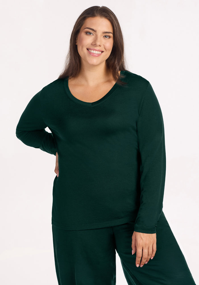 A woman smiles wearing a deep pine Woolx Lily V Neck Lounge Top, a long-sleeved Merino wool sleep shirt. 