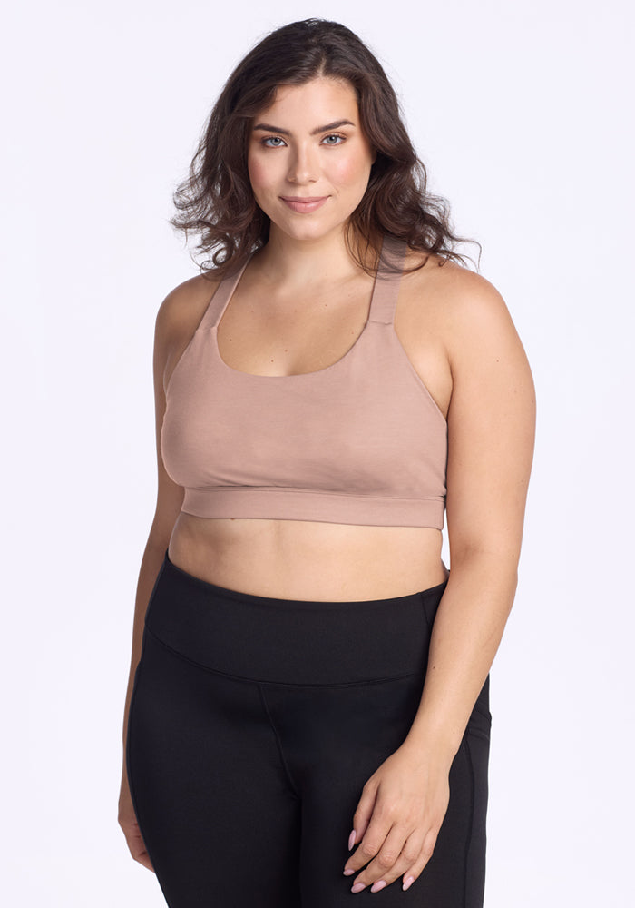 A woman with wavy brown hair smiles, wearing the Woolx Livi Bra in Morning Latte and black leggings, standing against a plain white background. 