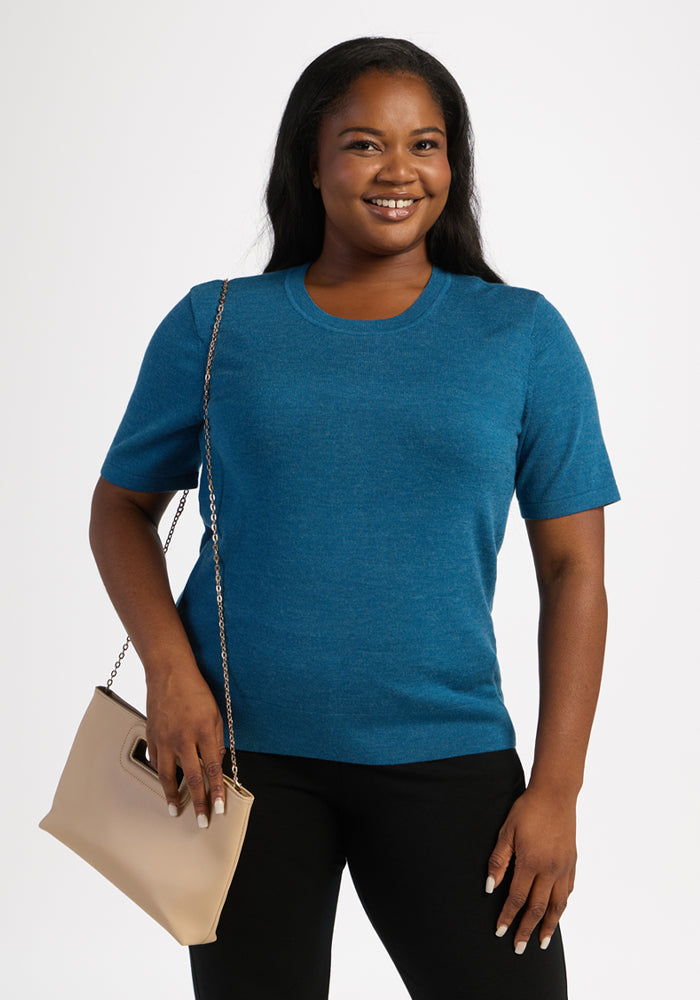 Woman in a blue Woolx Anna Crew Sweater holds a beige purse with chain strap, standing against a white background. 