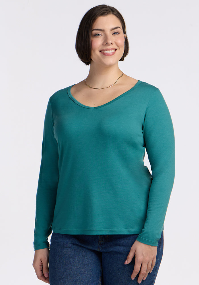 A woman with short brown hair smiles in a Woolx Layla V Neck Top - Surfside and blue jeans, standing against a plain white background. 