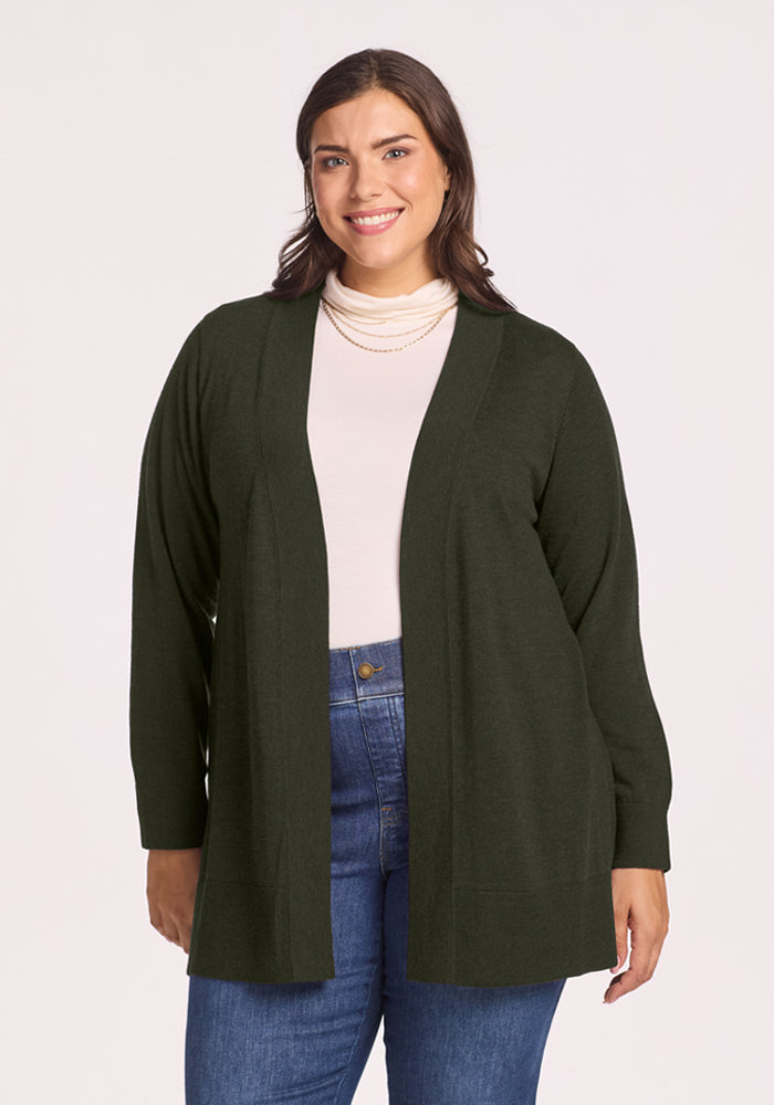 Woman in Woolx Macie Sweater - Duffel Bag, over a white top and blue jeans, smiling at camera against plain background. 