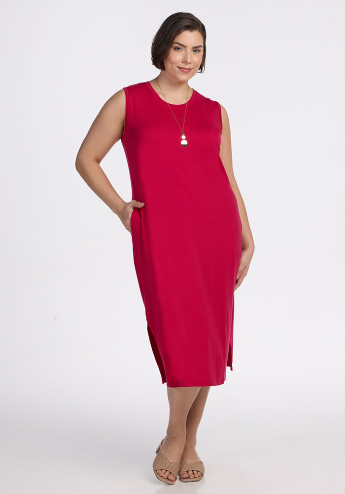 A woman with short brown hair models the Woolx Cassie Dress in Poppy Spark, a sleeveless, knee-length merino wool tank dress with side slits. She smiles, one hand in her pocket, wearing a necklace and beige sandals against a white background. 