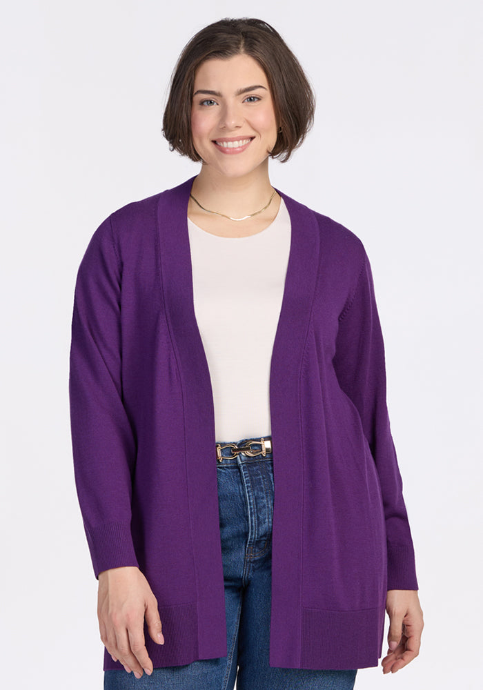 A woman with short brown hair smiles while wearing the Woolx Macie Sweater in Plum Crush over a white shirt and blue jeans, standing against a plain white background. 