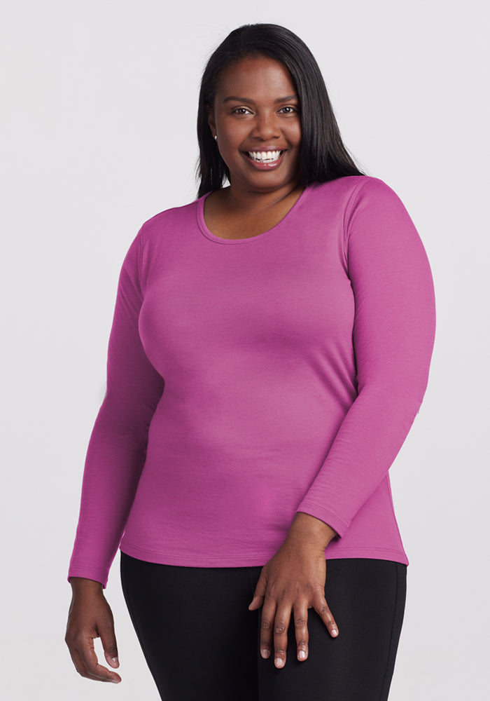 A woman with long dark hair smiles, wearing the Woolx Remi Long Sleeve T-Shirt in Purple Wine and black pants, standing against a plain light background. 