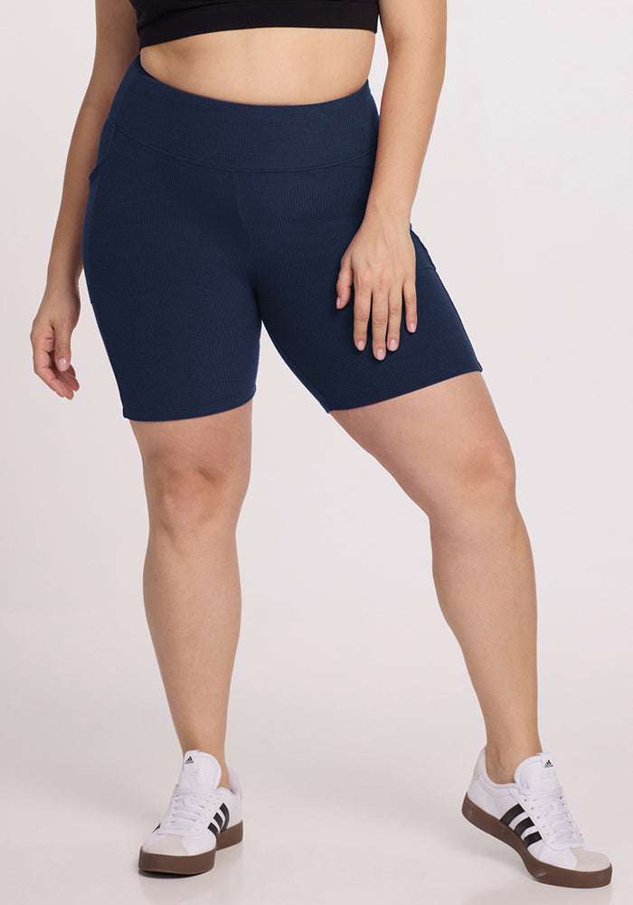 A person wearing a black crop top, Woolx Miley Bike Shorts in Deep Navy, and white Adidas sneakers stands against a light background with their left hand resting on their thigh. 