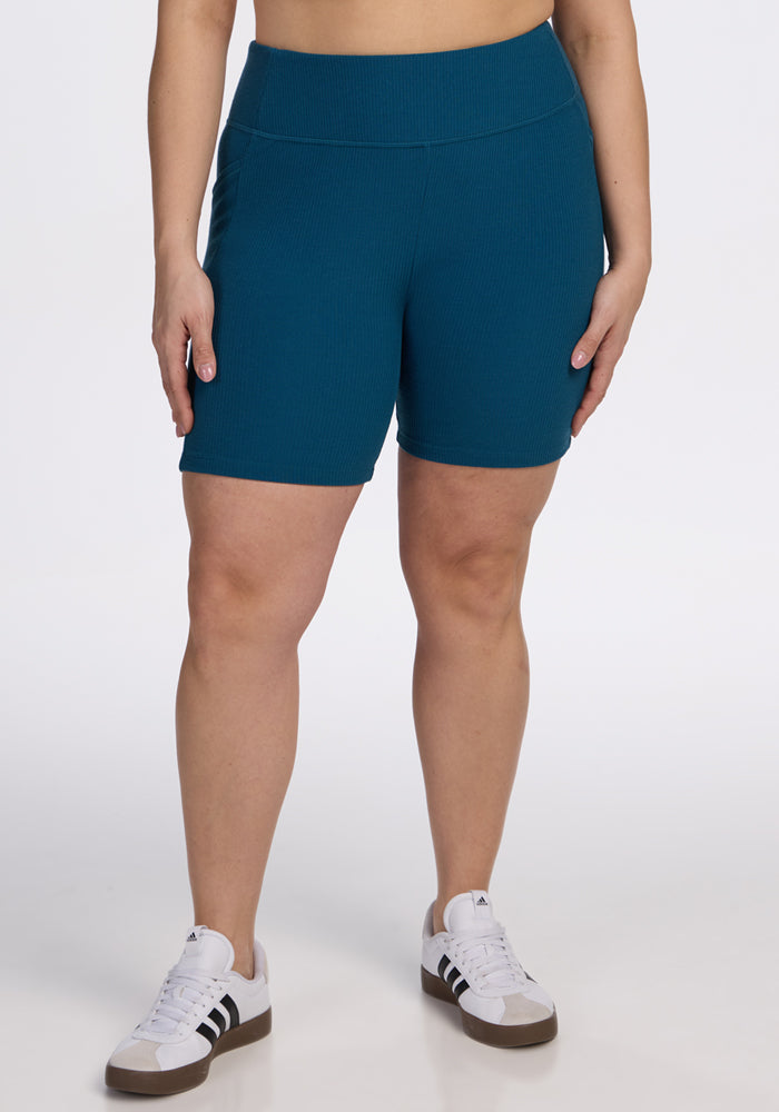 A person models the Woolx Miley Bike Shorts in Moroccan Blue, paired with white sneakers featuring black stripes, against a plain white background. Only their midsection and legs are shown. 