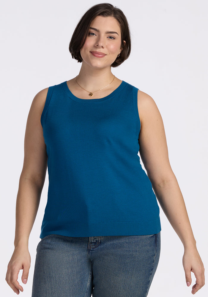 A woman with short brown hair, wearing the Woolx Chessy Tank Top in Moroccan Blue and blue jeans, stands against a plain white background, smiling gently at the camera. 