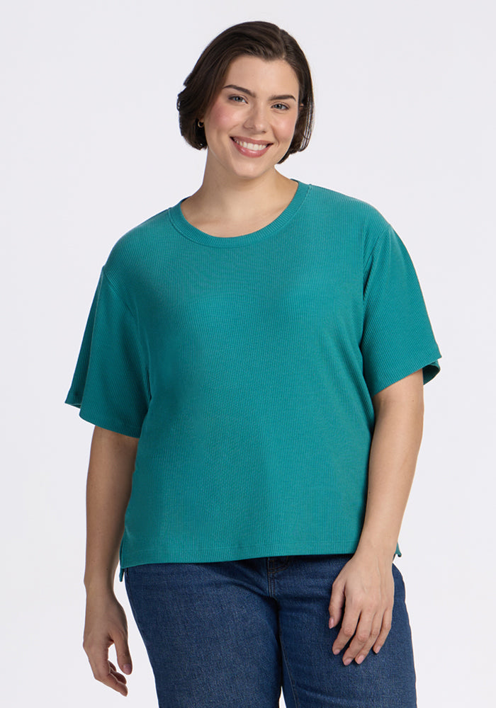 A woman with short brown hair, wearing the Woolx Delia Ribbed Short Sleeve - Surfside and blue jeans, stands smiling against a plain white background.