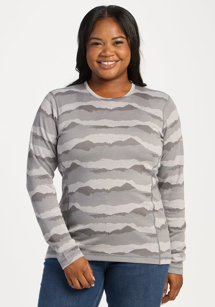 Smiling person wears Woolx Maddie Baselayer Top - High Peaks and blue jeans, standing against a white background. 