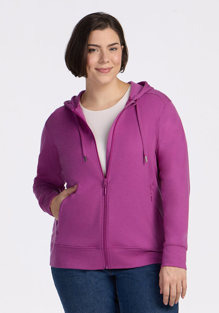 A woman with short brown hair smiles while wearing the Woolx Ryann Hoodie in Purple Wine over a white shirt and blue jeans, standing against a plain white background. 