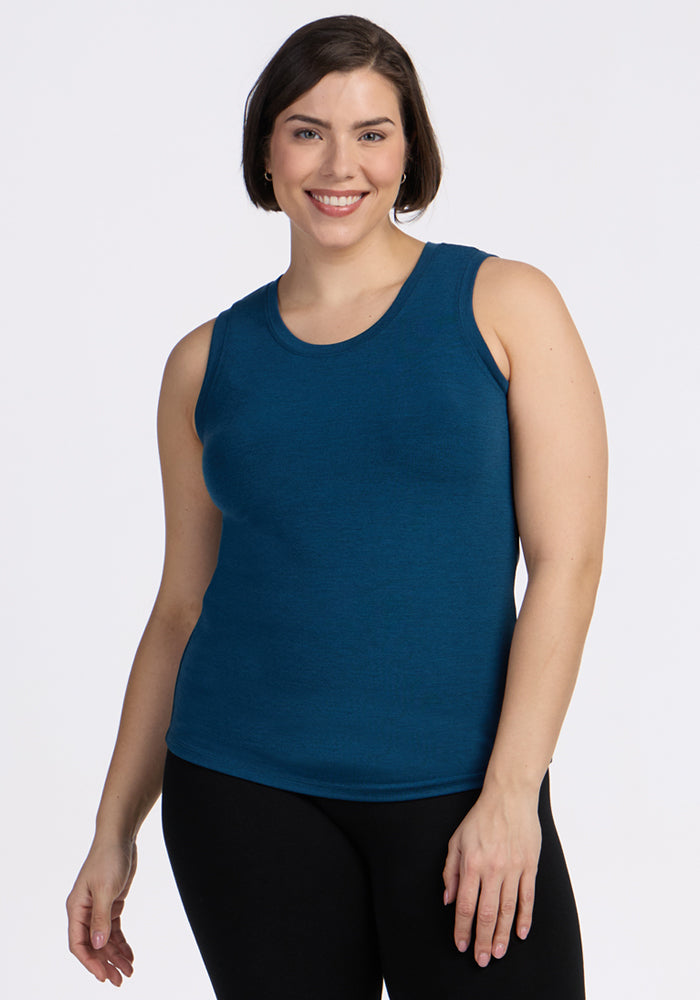 A woman with short brown hair smiles while wearing the Woolx Ava Tank Top in Majolica Blue and black pants, standing against a plain white background. 