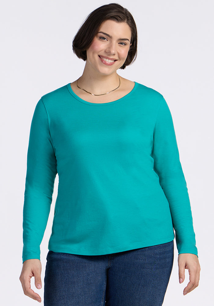 A woman with short brown hair is smiling and wearing the Woolx Remi Long Sleeve T-Shirt in Cool Jade with blue jeans, standing against a plain white background. 