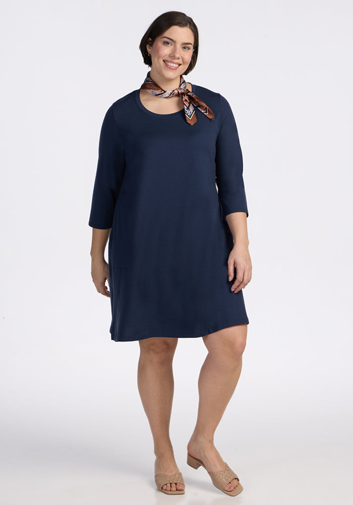 A woman smiles against a white background in the Woolx Rissa A Line Dress - Deep Navy, styled with a patterned scarf and beige open-toe shoes—an ideal look for fall and winter layering.