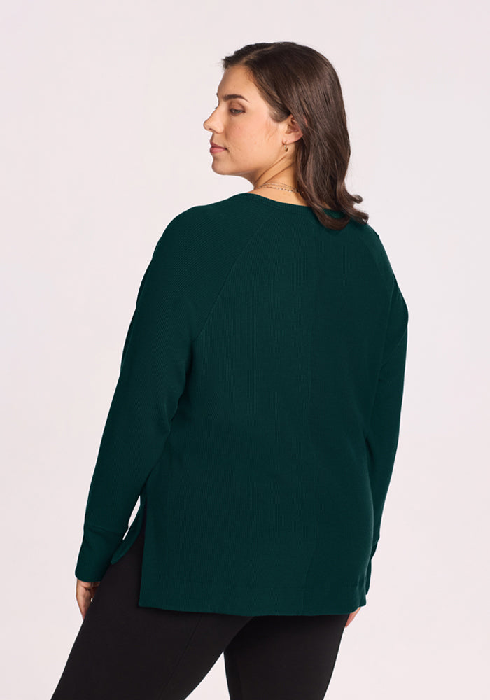 A woman with dark hair faces away, wearing the Woolx Ainsley Sweater in Deep Pine and black pants.