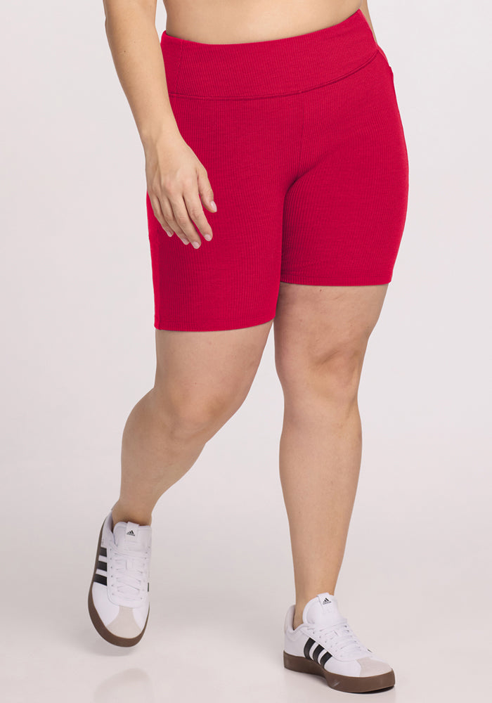 A person wearing Woolx Miley Bike Shorts in Poppy Spark and white Adidas sneakers with black stripes stands against a plain light background, cropped from the midriff down. 