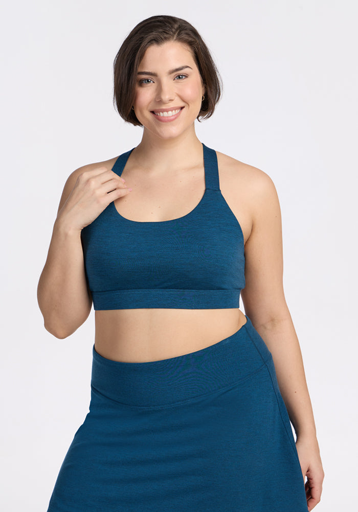 A woman with short brown hair smiles, wearing the Woolx Livi Bra in Majolica Blue, paired with a matching skirt, standing against a white background. 