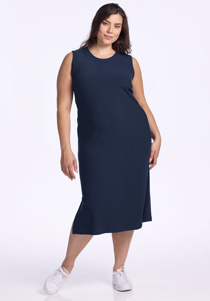 A woman with long brown hair wears the Woolx Iris Ribbed Dress in Deep Navy and white sneakers, standing against a plain white background and smiling slightly. 