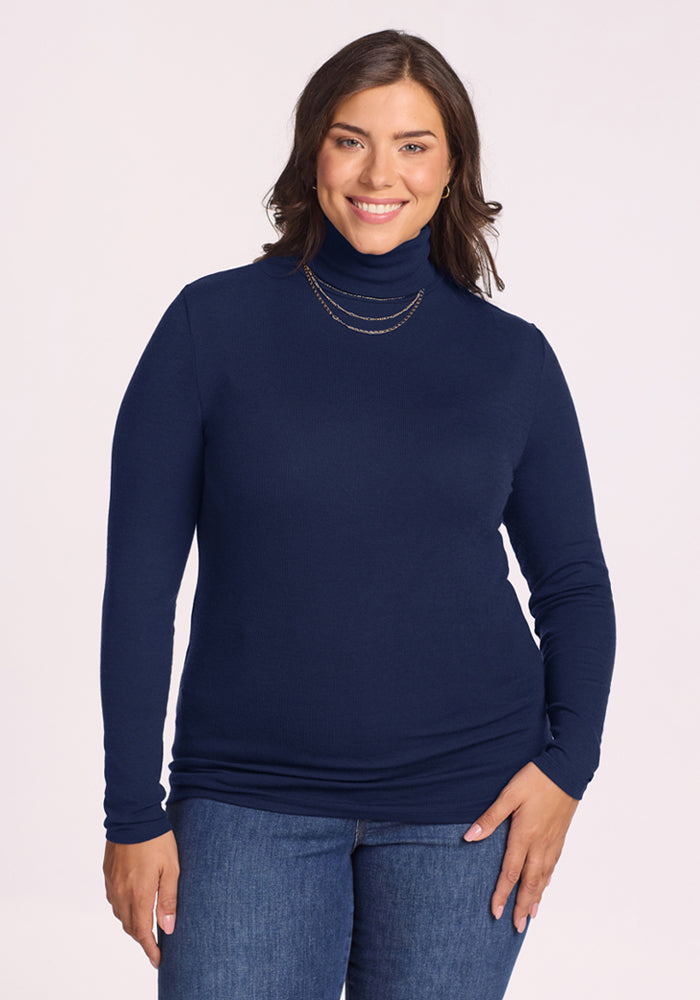 Smiling woman in Woolx Sage Ribbed Turtleneck—Ocean Cavern, blue jeans, and gold necklace, facing the camera. 