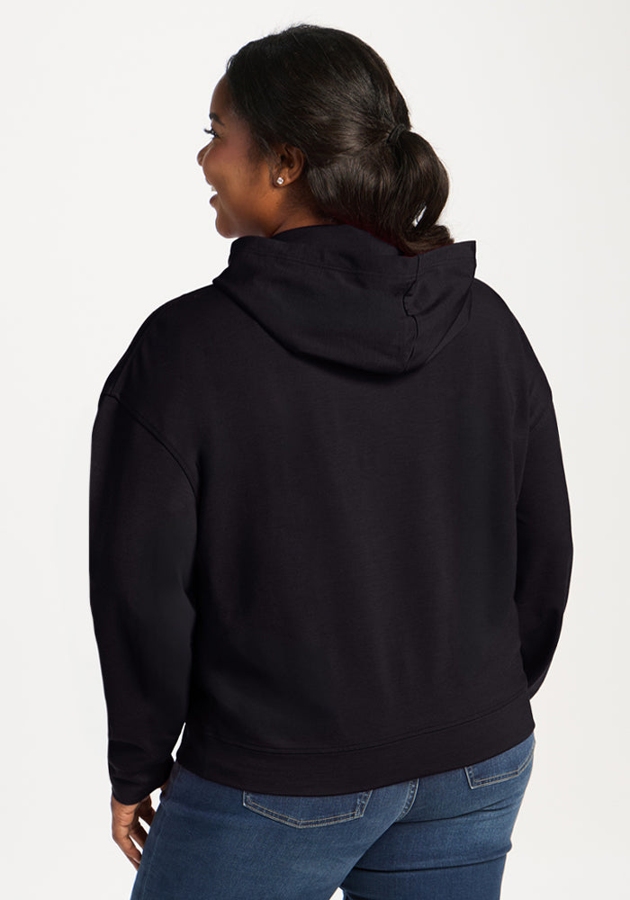 A woman in the Woolx Lizzy Hoodie - Black and blue jeans stands facing away, looking to the side, on a white background.
