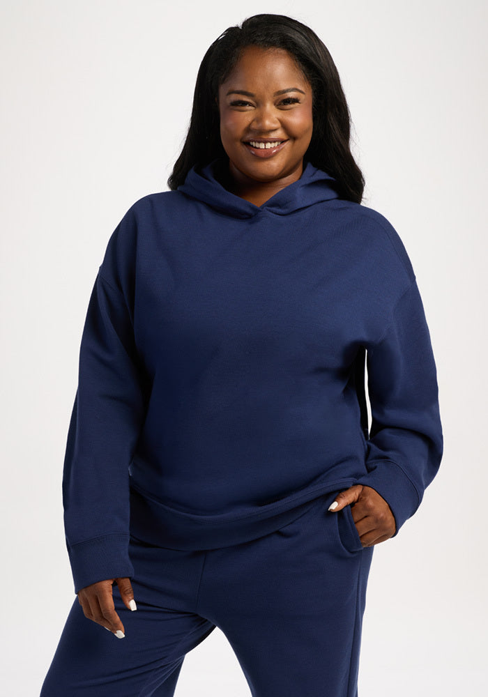 A smiling woman wearing a dark blue Genevieve Hoodie - Ocean Cavern featuring moisture-wicking fabric and a front pocket. 