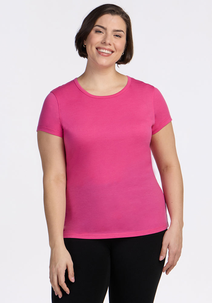 A person with short brown hair smiles against a white background, wearing the Woolx Addie Short Sleeve Crew in Raspberry Fizz—an odor-free, lightweight Merino wool t-shirt—paired with black pants. 