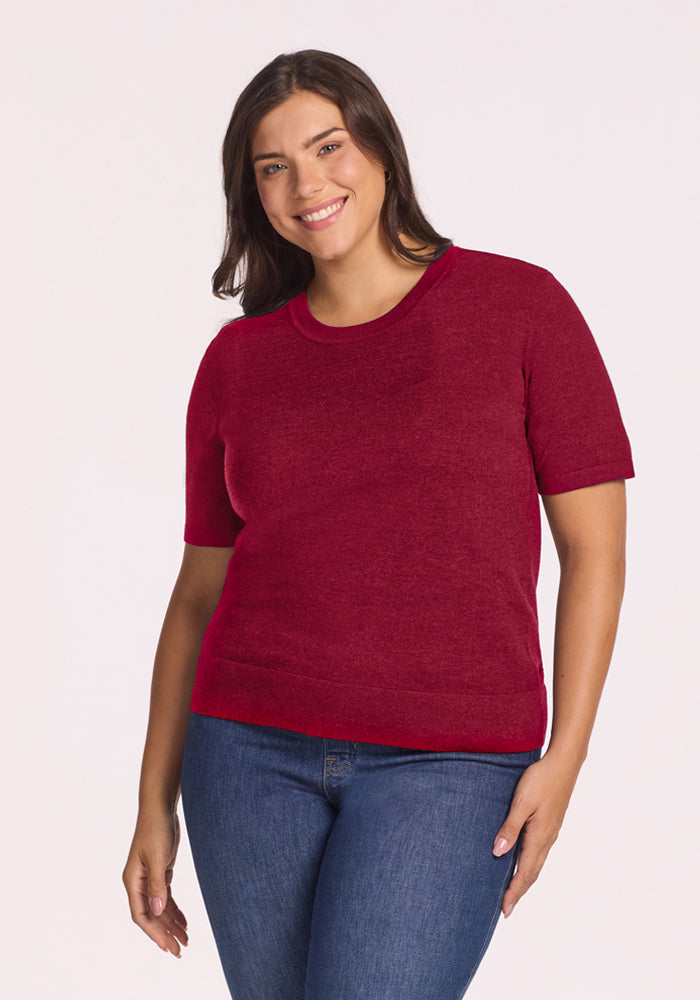 Woman smiles in a Beet Red Anna Crew Sweater by Woolx and blue jeans, against a plain light background. 