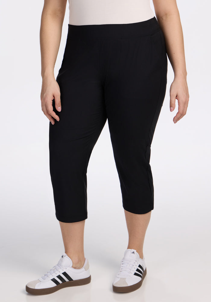 A person wearing Woolx Nicole Crop Pants in black, a white top, and white Adidas sneakers with black stripes stands against a plain white background. 