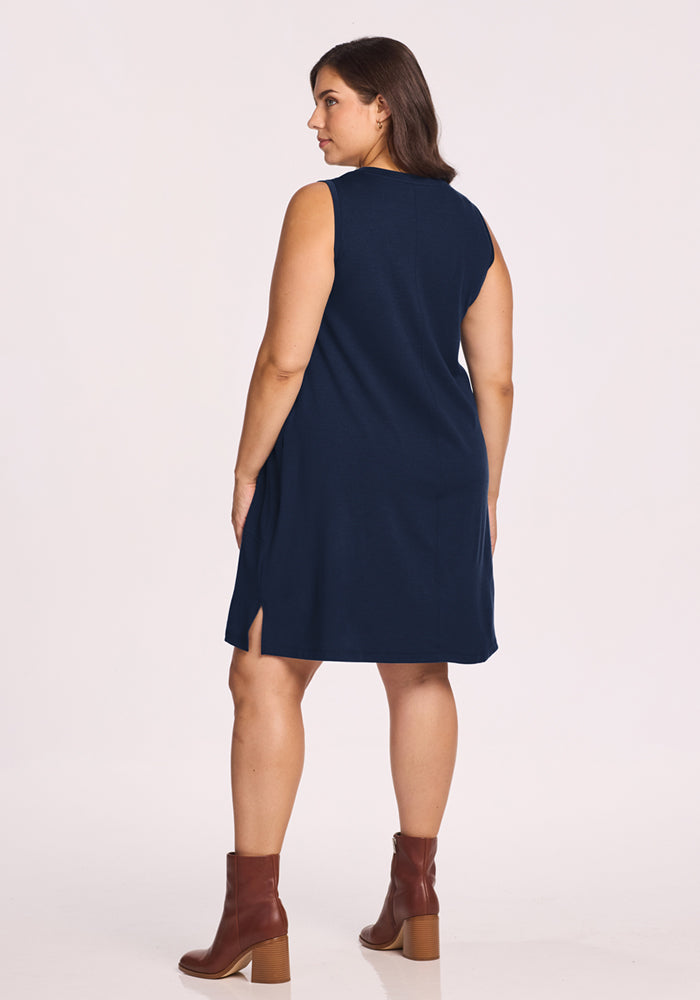 A woman faces away, wearing the Woolx Clara Dress in Deep Navy—it's sleeveless, breathable, with small side slits—and brown ankle boots. The background is plain and light-colored.