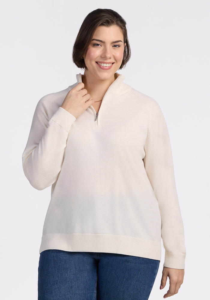 A woman with short brown hair smiles at the camera, wearing Woolx's Collins Half Zip Sweater in Dewdrop Pearl and blue jeans against a plain white background. 