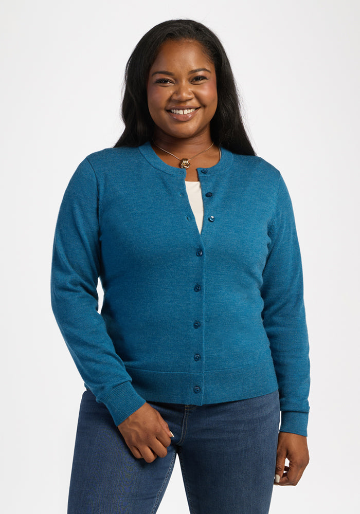 Woman smiling in a Woolx Caitlin Sweater - Blue Pine over a light top and jeans, standing against a plain background. 
