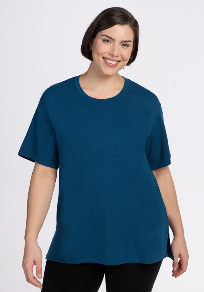 A smiling woman with short dark hair wears the Woolx Audrey Tee in Majolica Blue and black pants, standing against a plain white background. 