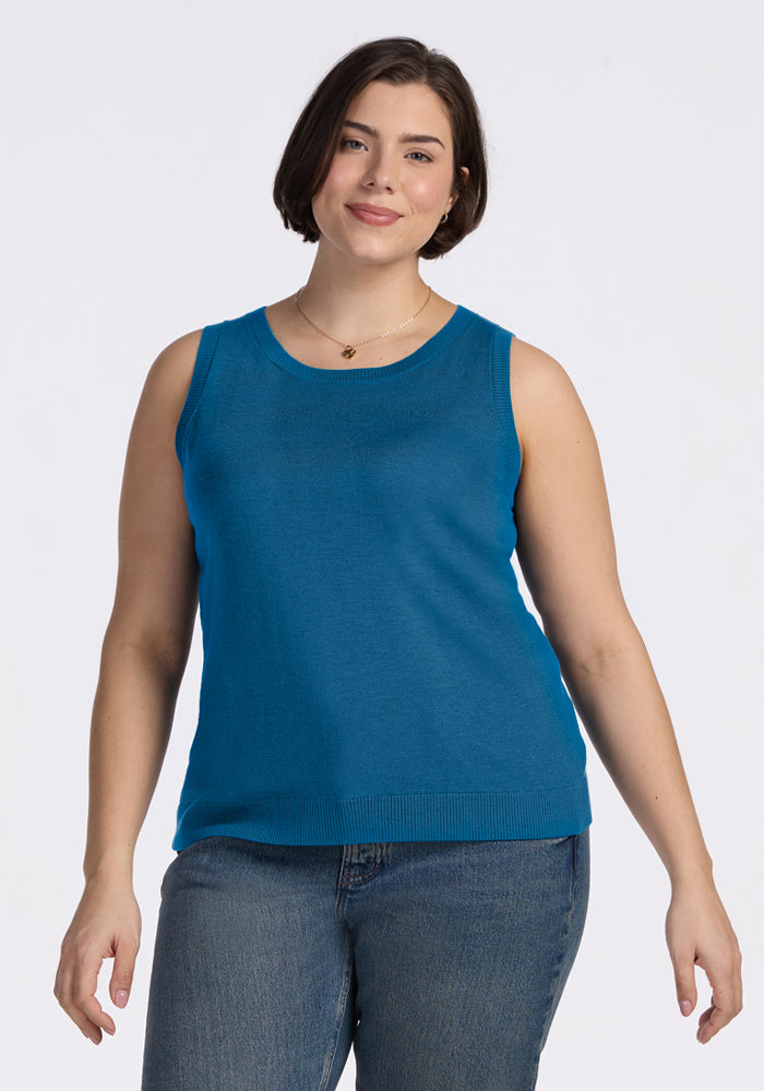 Woman in a Woolx Chessy Tank Top - Blue Pine and jeans stands against a white background, smiling gently. 