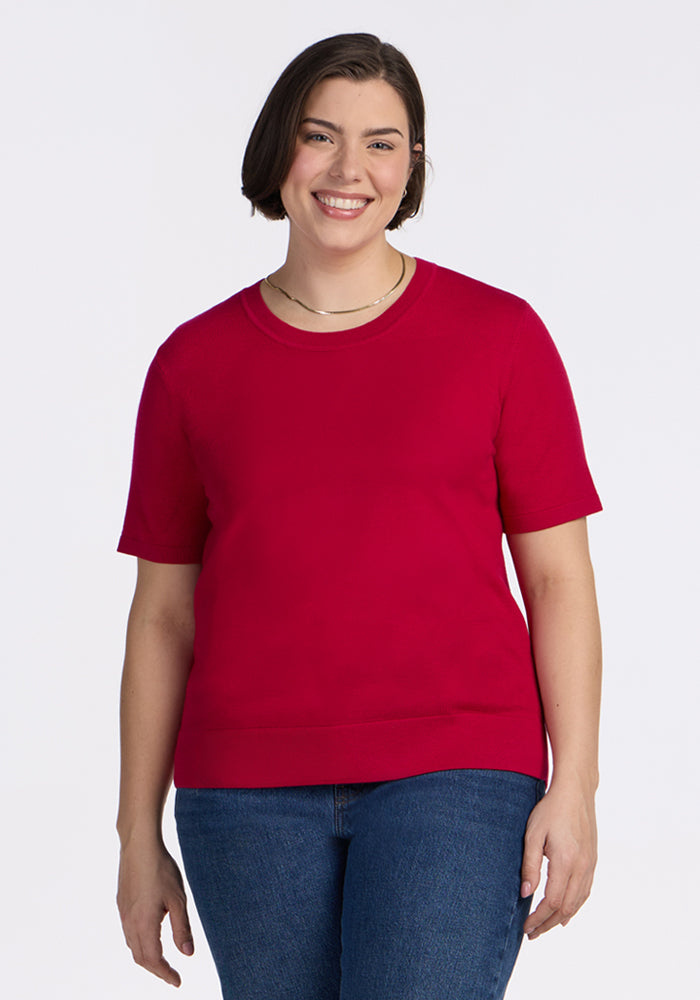 A woman with short brown hair smiles against a white background, wearing the Woolx Anna Crew Sweater in Poppy Spark—a chic short-sleeve style—paired with classic blue jeans. 
