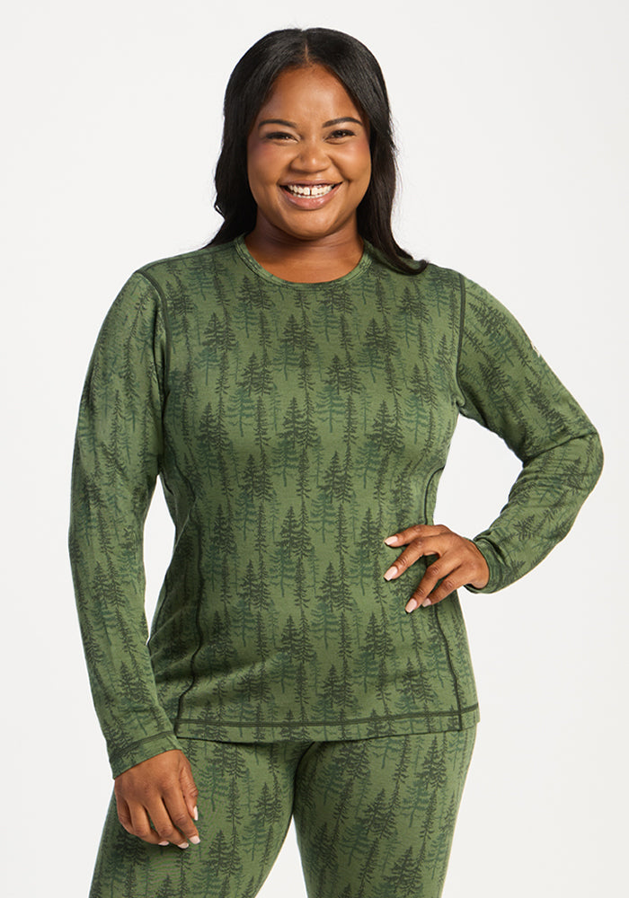 Woman smiles, wearing Woolx Maddie Baselayer Top - Forest Friends, hand on hip in green, dark tree pattern set. 