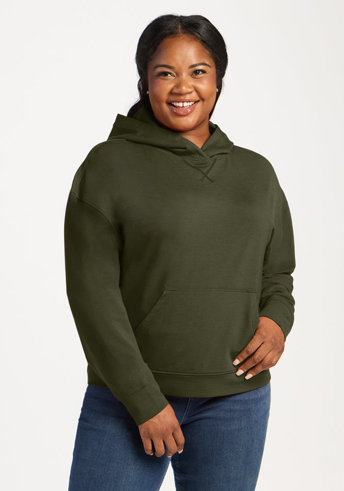 Smiling woman in Woolx Lizzy Hoodie - Everleaf and blue jeans stands against a plain white background.