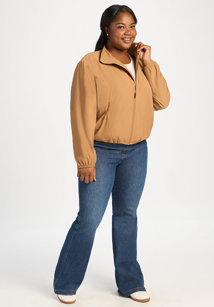 A smiling woman wears a tan, high-collared Woolx Cooper Windbreaker - Saddleback and blue jeans.