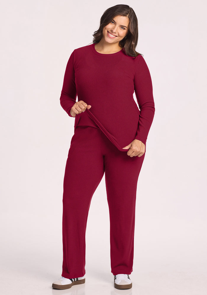Smiling woman in Woolx Hadley Ribbed Crew - Beet Red, holding the hem, stands on a white background.