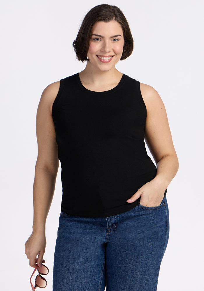 A woman with short brown hair, wearing the Woolx Ava Tank Top in black and blue jeans, smiles at the camera against a plain white background, holding sunglasses in her left hand. 