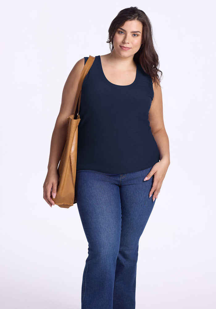 A woman with long dark hair smiles slightly as she stands against a plain white background, wearing the Woolx Sloane Ribbed Tank in Deep Navy and blue jeans, and holding a tan shoulder bag.