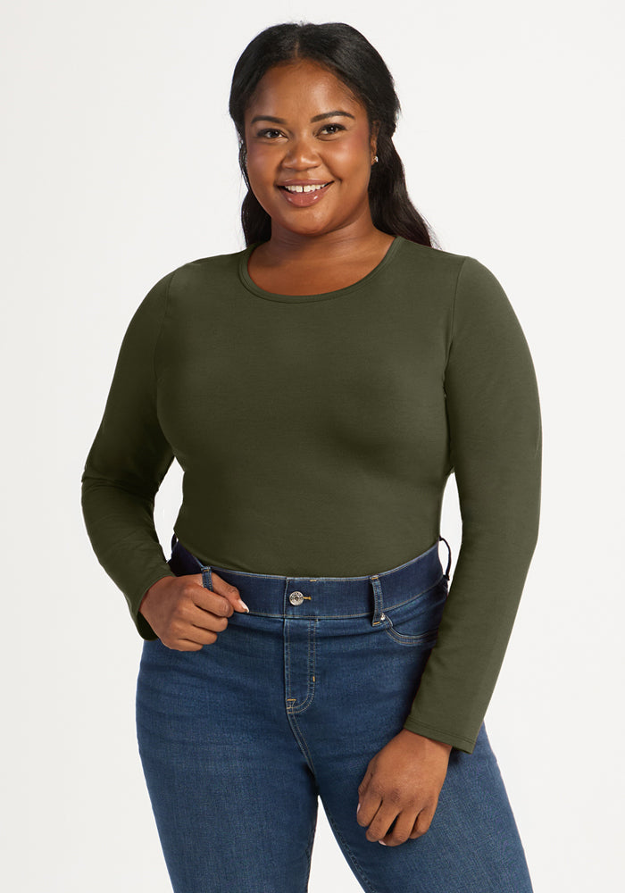 Woman in the Woolx Margot Bodysuit in Everleaf and blue jeans smiles against a plain white background. 