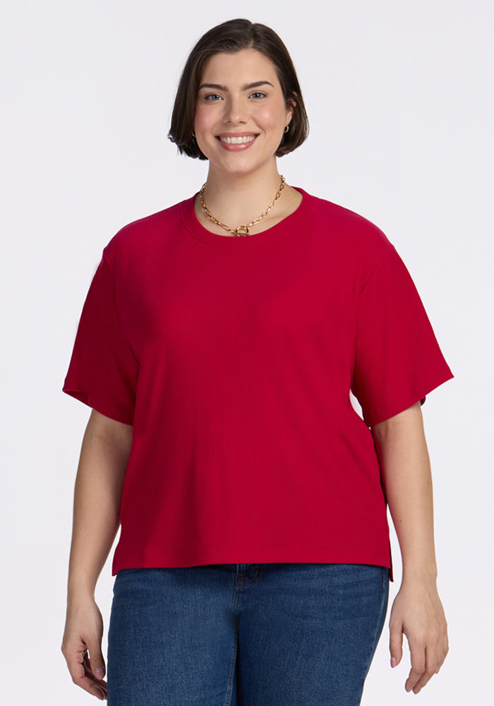 A woman with short brown hair, smiling against a plain white background, wears the Woolx Delia Ribbed Short Sleeve in Poppy Spark, paired with blue jeans and a gold necklace. 