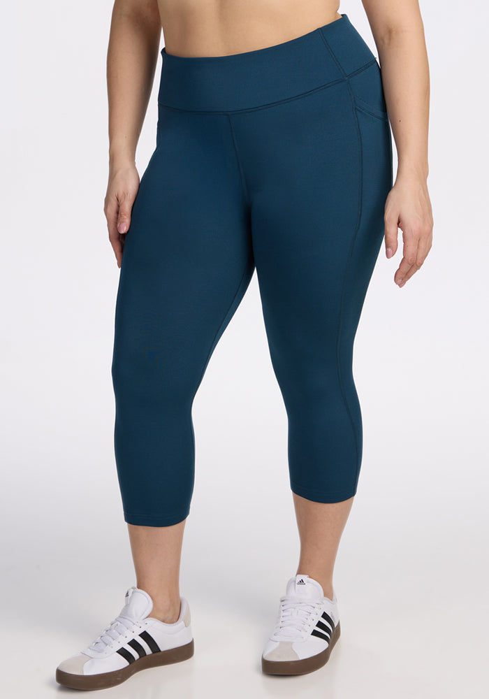 A person stands against a white background wearing Woolx Merino Flex® McKenna Capris in Majolica Blue and white Adidas sneakers with black stripes. The image is cropped waist to ankles. 