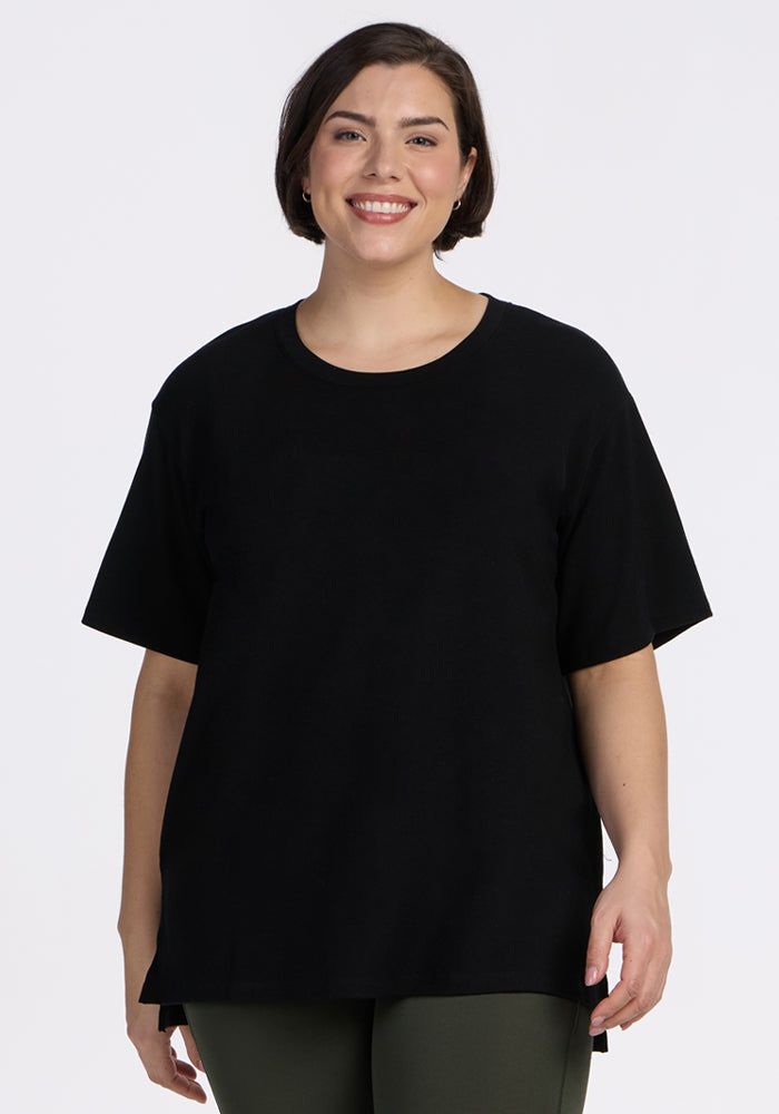 A woman with short brown hair smiles while wearing the Woolx Audrey Tee in black and dark green pants, standing against a plain white background. 