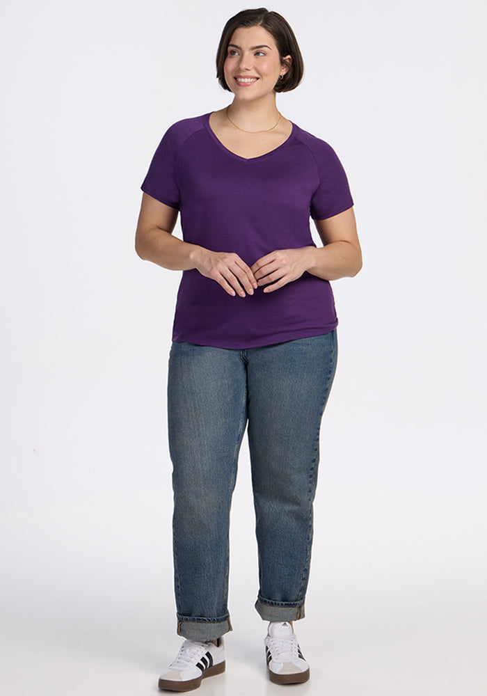A woman with short brown hair, wearing the Woolx Mia V Neck - Plum Crush shirt, blue jeans, and white sneakers, stands smiling against a plain white background.
