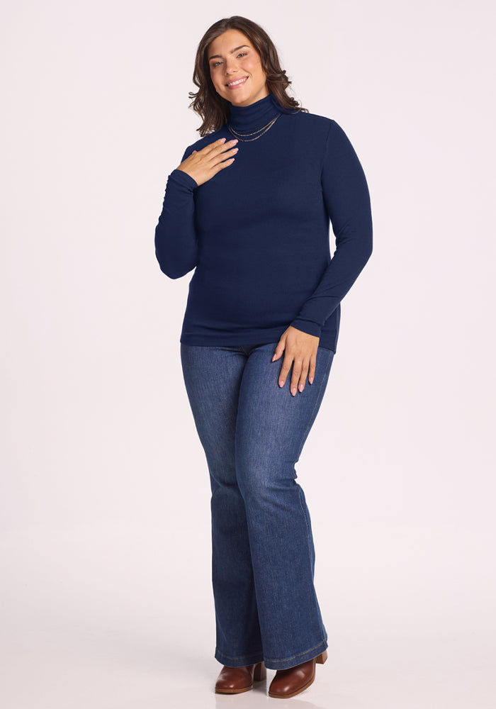 Smiling woman wears the Woolx Sage Ribbed Turtleneck in Ocean Cavern, blue jeans, and brown boots against a white background.