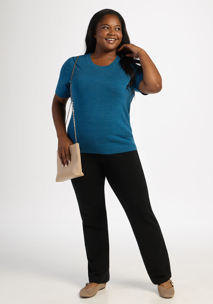 A woman smiles in the Woolx Anna Crew Sweater - Blue Pine, black pants, beige shoes, and holds a beige purse on her shoulder.